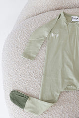 Bailey Onesie in Desert Sage and Clover laid on a cream chair, showing fine ribbed fabric, long sleeves, a front zip, hny embroidery and a dark foot cuff.