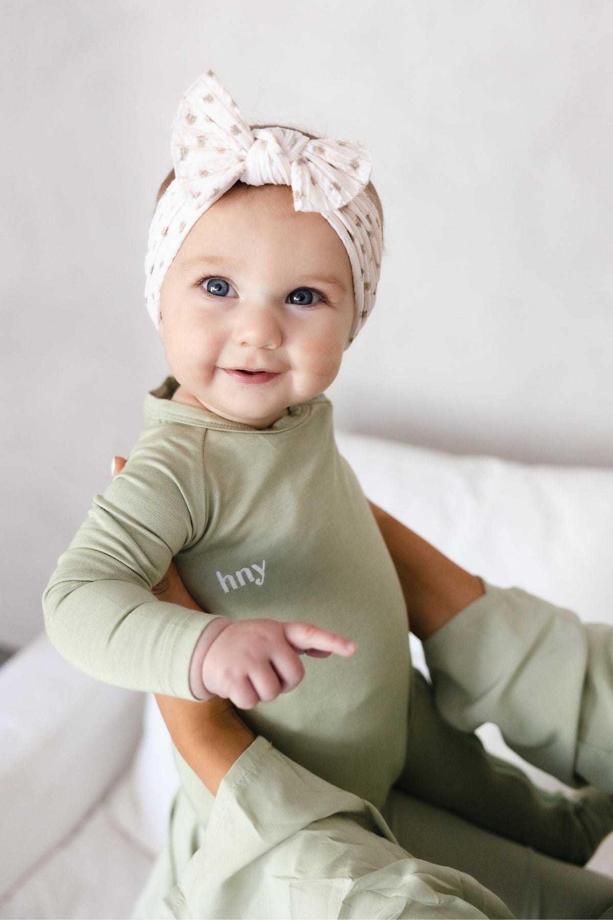 Close-up of a smiling baby supported by an adult, wearing the sage green Bailey Onesie with long sleeves, a round neckline and white hny chest embroidery.