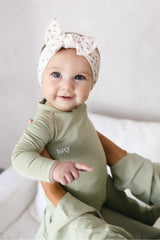 Close-up of a smiling baby supported by an adult, wearing the sage green Bailey Onesie with long sleeves, a round neckline and white hny chest embroidery.